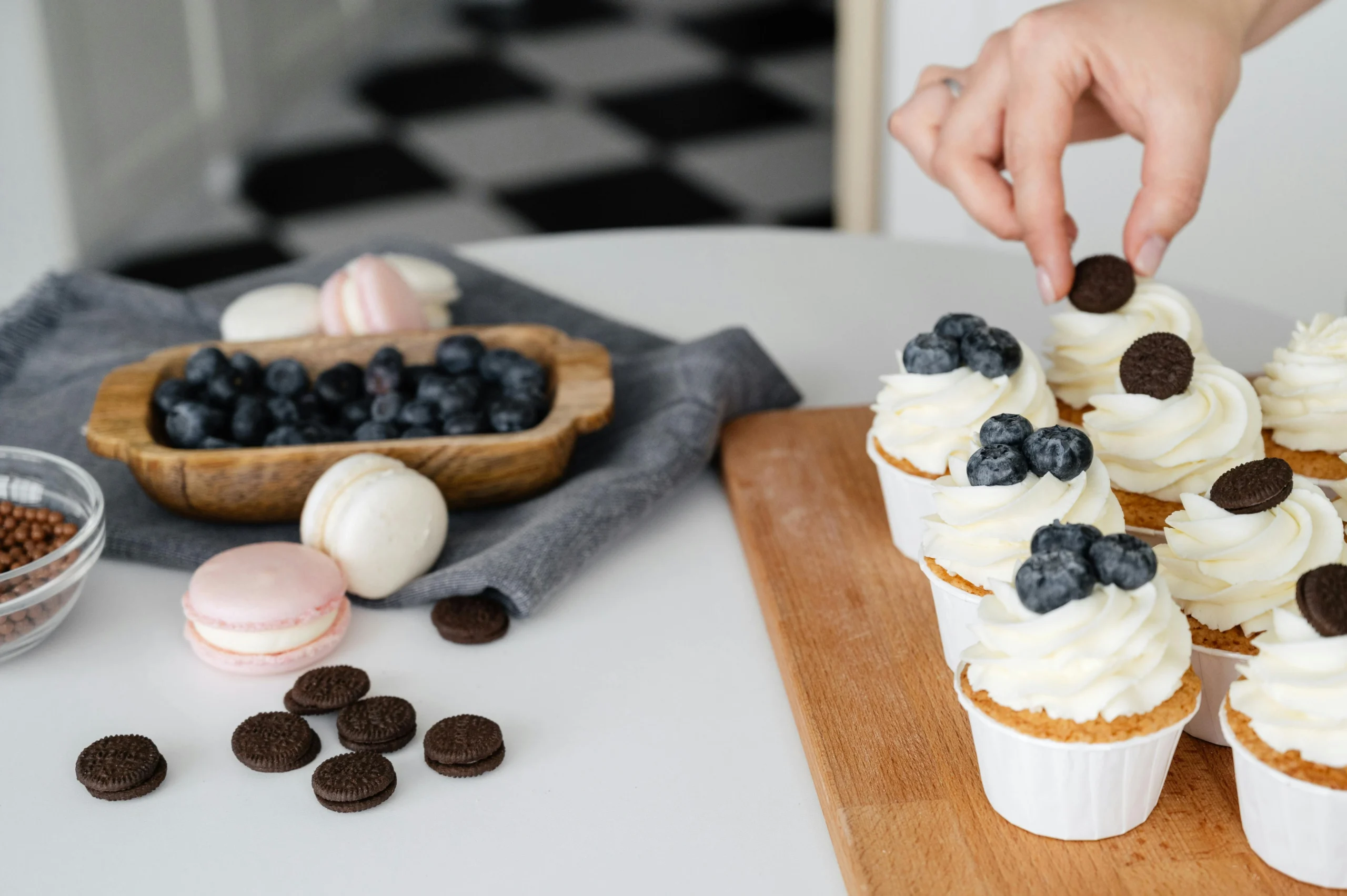 Hand som dekorerar cupcakes med vitchokladfrosting och blåbär på träbricka, med macarons och Oreo-kakor i förgrunden