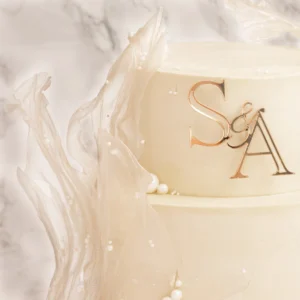 Närbild på bröllopstårtas guldmonogram S&A omgivet av isochokladsegeldekor och vita pärlor