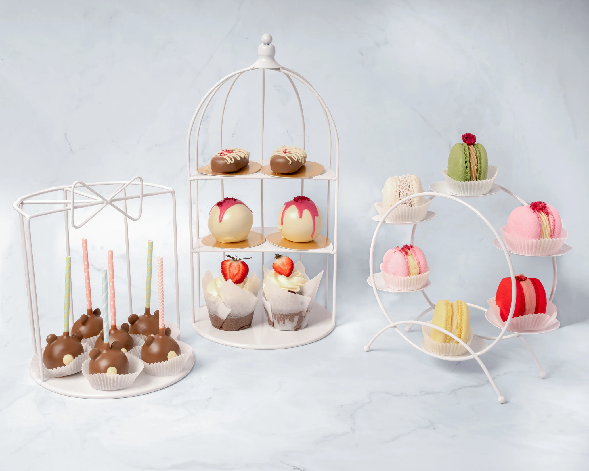 Tre vita dekorativa ställningar med cake pops, popsicle cakes och macarons på marmorbord