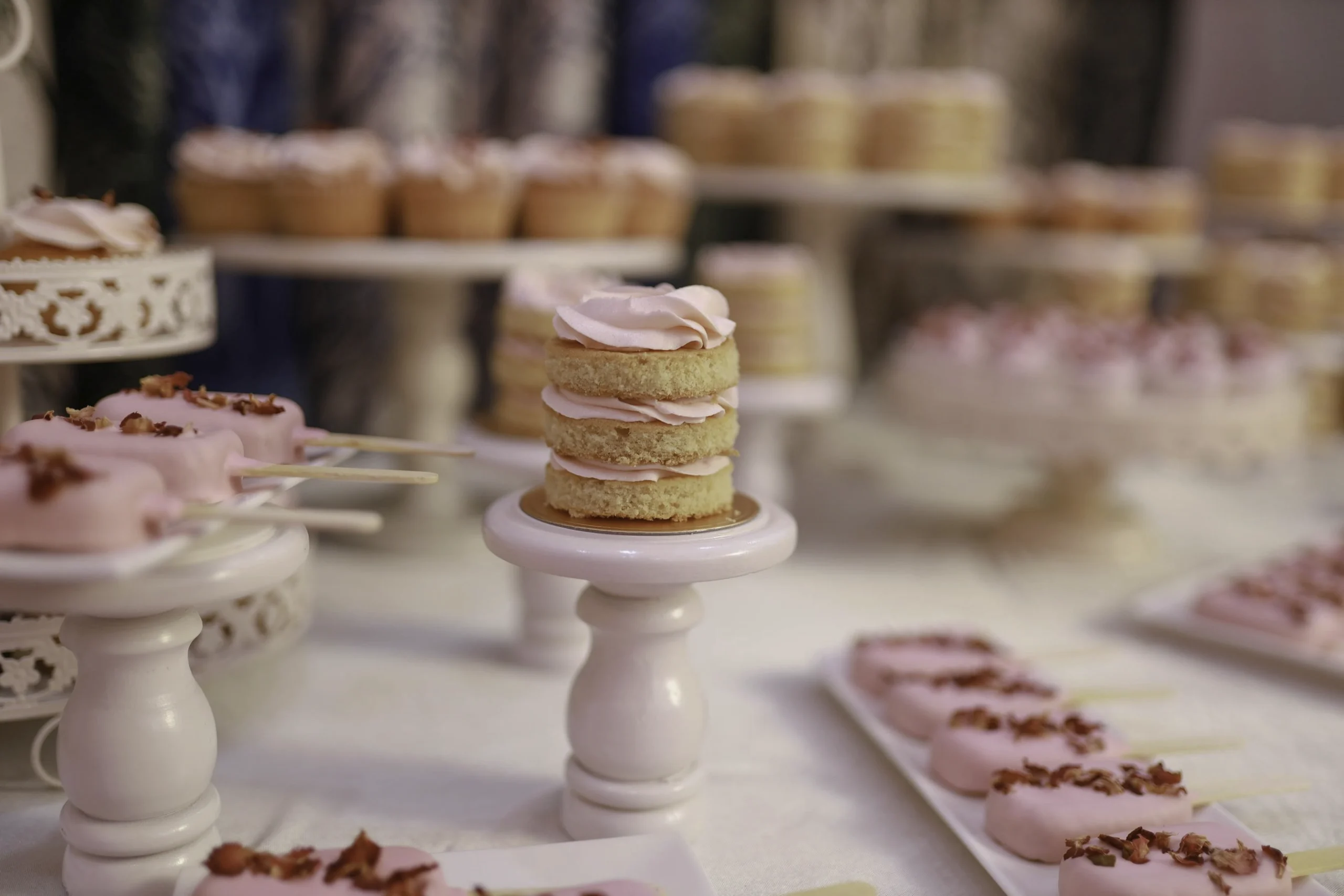 Elegant candy bar-uppställning med minilagertårta med rosa frosting på vit piedestal, rosa chokladdoppade popsicles med torkade rosenblad och cupcakes på vita tårtfat