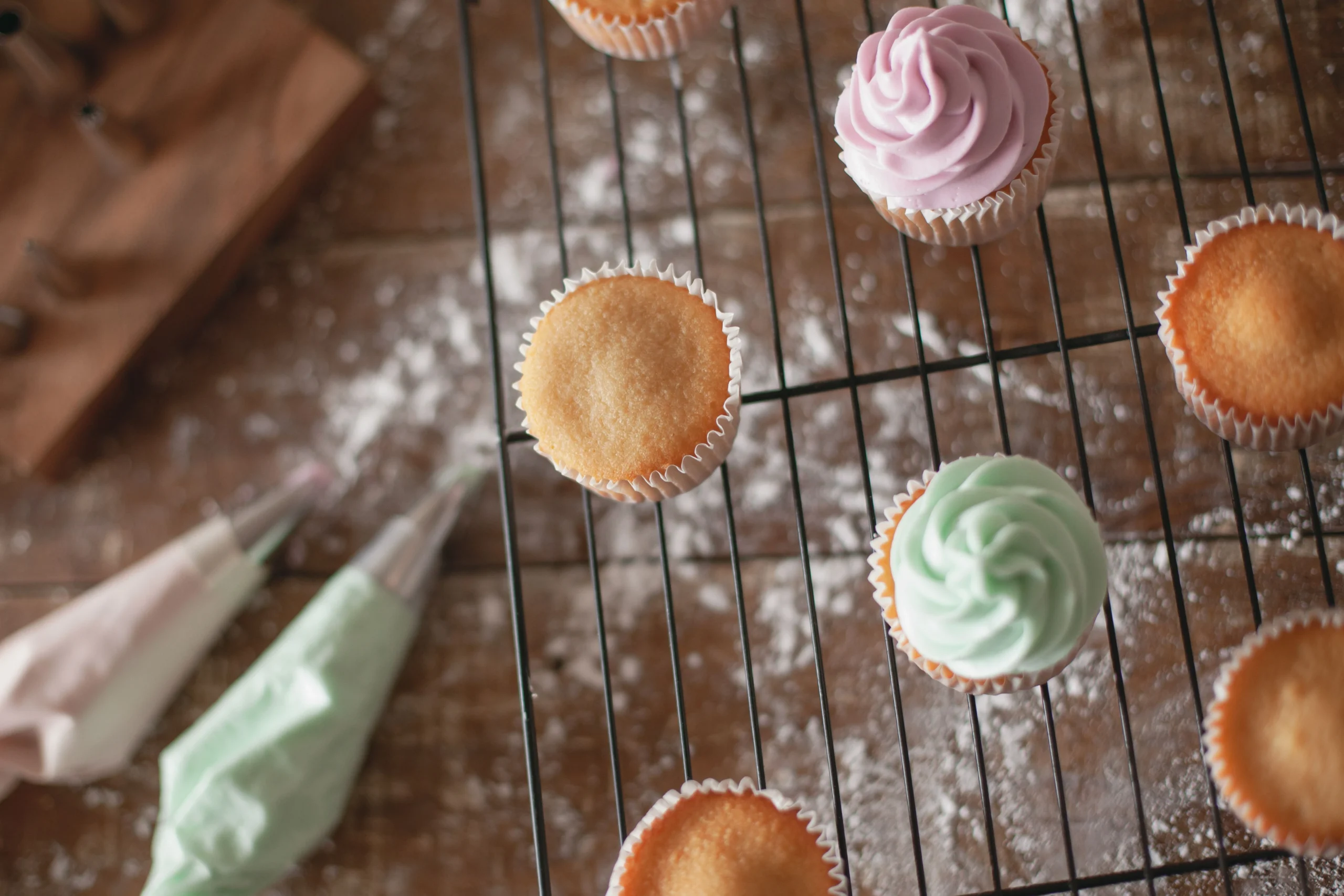 Uppifrånvy av vaniljcupcakes på bakgaller, några utan och några med rosa och mintgrön frosting, med spritspåsar i förgrunden på mjölbestänkt träbord