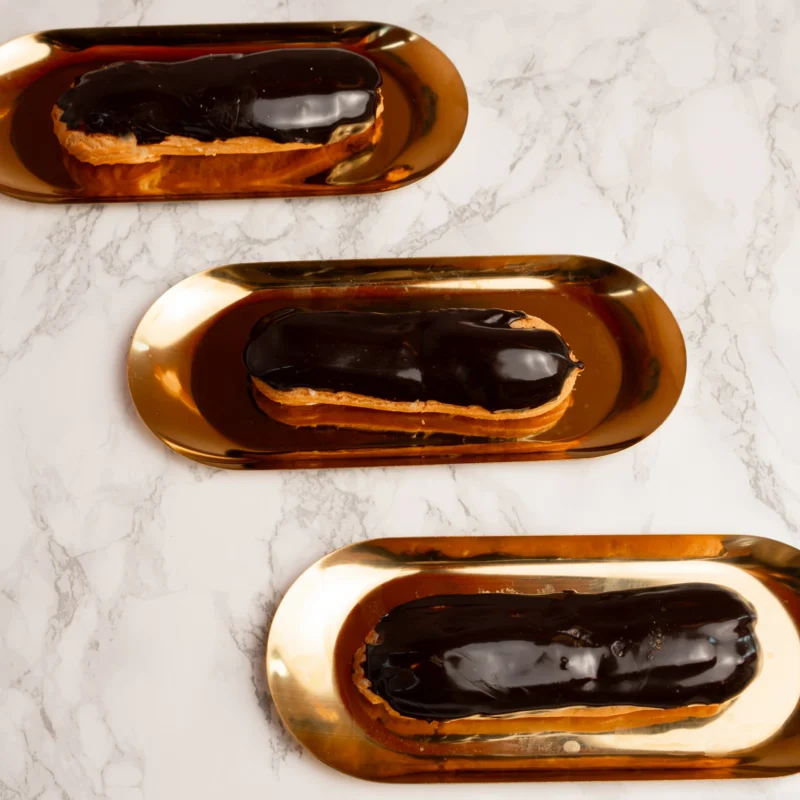 Tre chokladglaçerade eclairer på guldfärgade serveringsbrickor fotograferade ovanifrån mot marmorbakgrund
