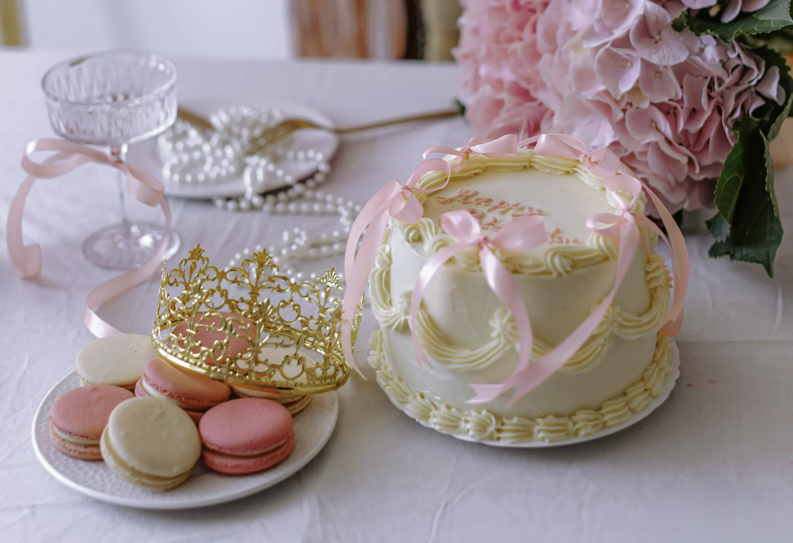 Vit gräddborderad födelsedagstårta med rosa sidenband och rosetter, bredvid tallrik med rosa och beige macarons och guldkrona på vitt bord med hortensiabukett