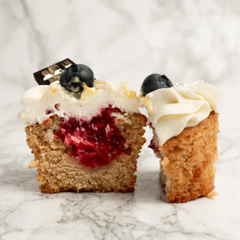 Ljus cupcake delad på mitten med synlig röd hallonfyllning, vispgrädde, blåbär och chokladbit på toppen