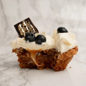 Ljus cupcake delad på mitten med synlig karamellfyllning, vispgrädde, blåbär och chokladbit på toppen