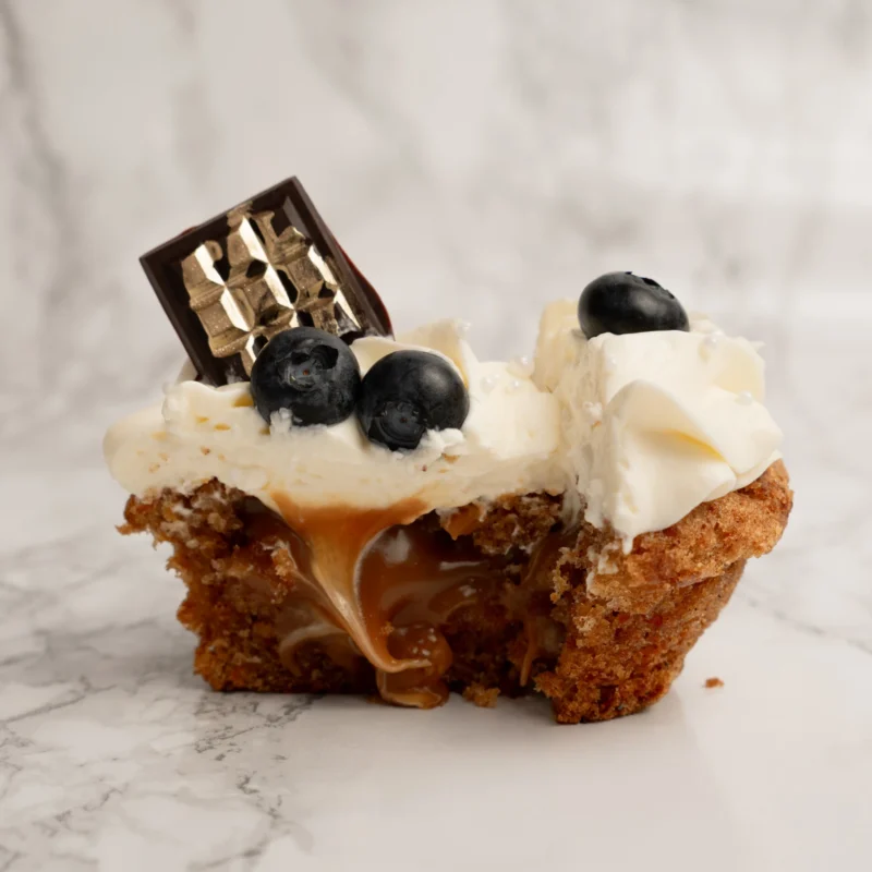 Ljus cupcake delad på mitten med synlig karamellfyllning, vispgrädde, blåbär och chokladbit på toppen