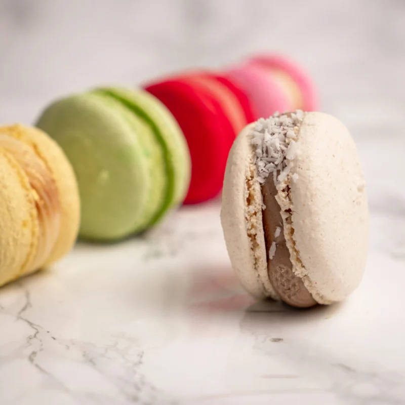 Närbild på beige kokos macaron i fokus med övriga färgglada macarons suddiga i bakgrunden