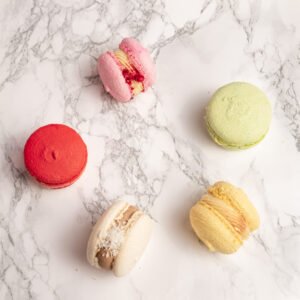 Fem macarons i olika färger spridda ovanifrån mot marmorbakgrund: rosa, grön, röd, beige och gul