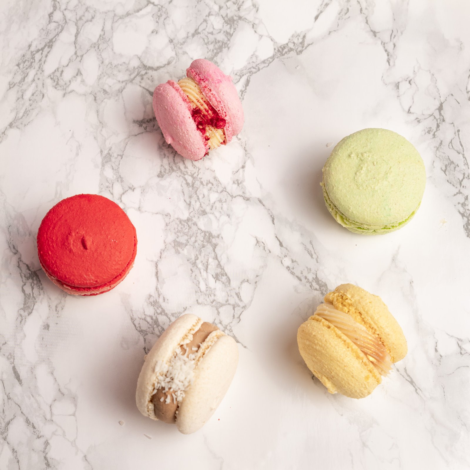 Fem macarons i olika färger spridda ovanifrån mot marmorbakgrund: rosa, grön, röd, beige och gul