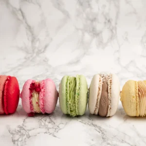 Fem macarons i rad framifrån i färgerna röd, rosa, grön, beige och gul mot marmorbakgrund