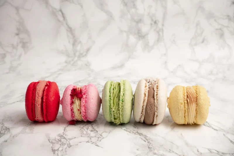 Fem macarons i rad framifrån i färgerna röd, rosa, grön, beige och gul mot marmorbakgrund