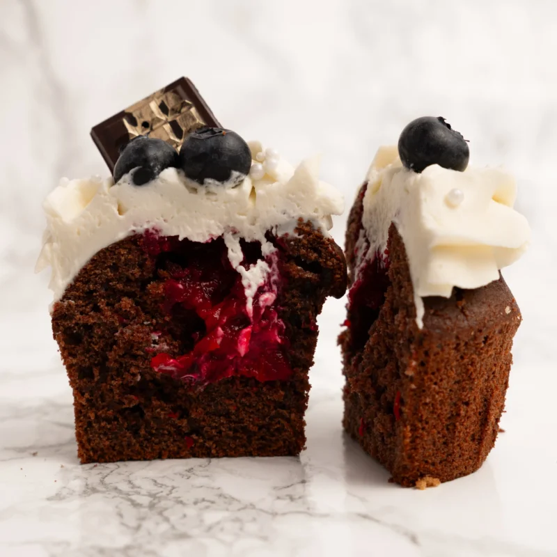 Mörk cupcake delad på mitten med synlig röd hallonfyllning, vispgrädde, blåbär och chokladbit på toppen