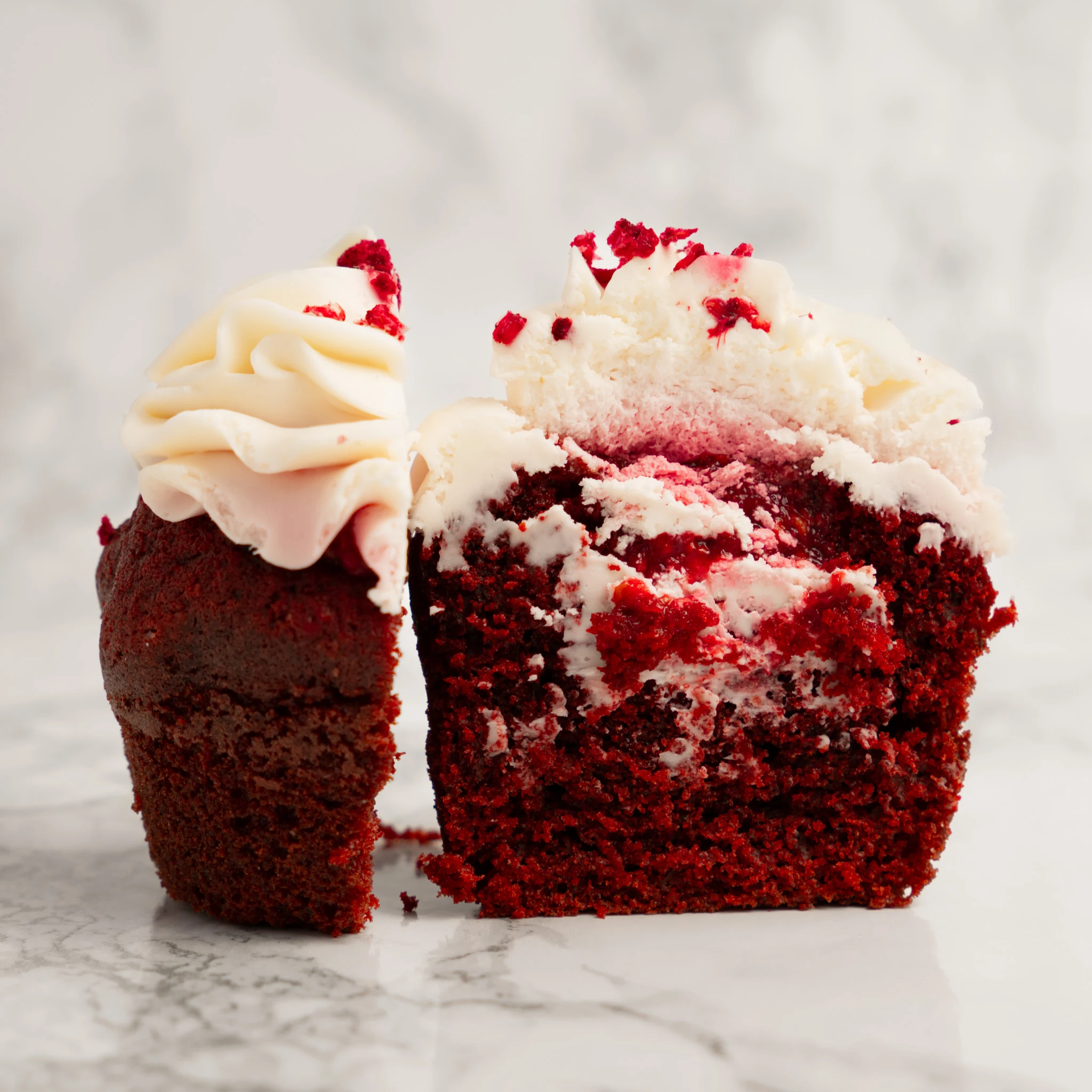 Red Velvet cupcake delad på mitten med synlig röd deg och cream cheese-fyllning, toppat med cream cheese-frosting och torkade hallonbitar