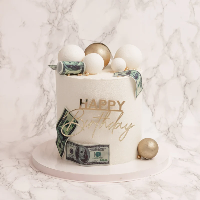 Spheres & Dollars tårta framifrån med vita och guldsfärer, dollarsedlar och Happy Birthday-topper mot marmorbakgrund