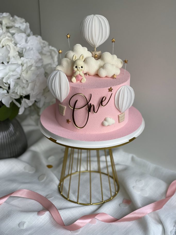 Tårta i rosa med moln och ballonger, dekorerad med figurer för ett första födelsedagsfirande.