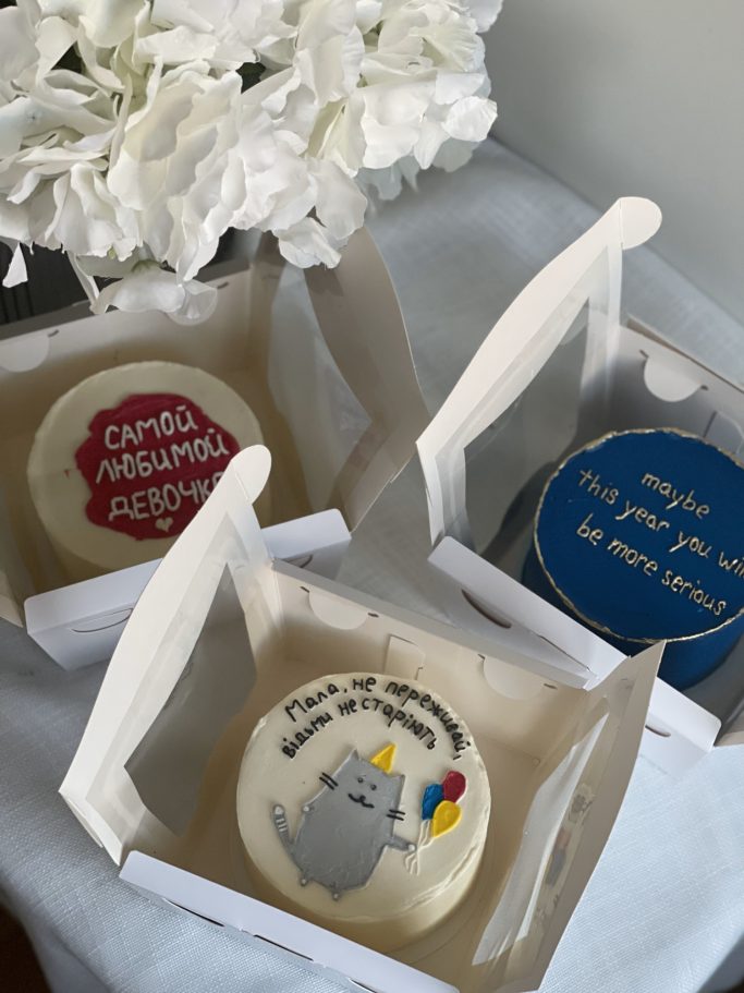 Tre cupcakes i papplådor med olika färgglada dekorationer och texter.